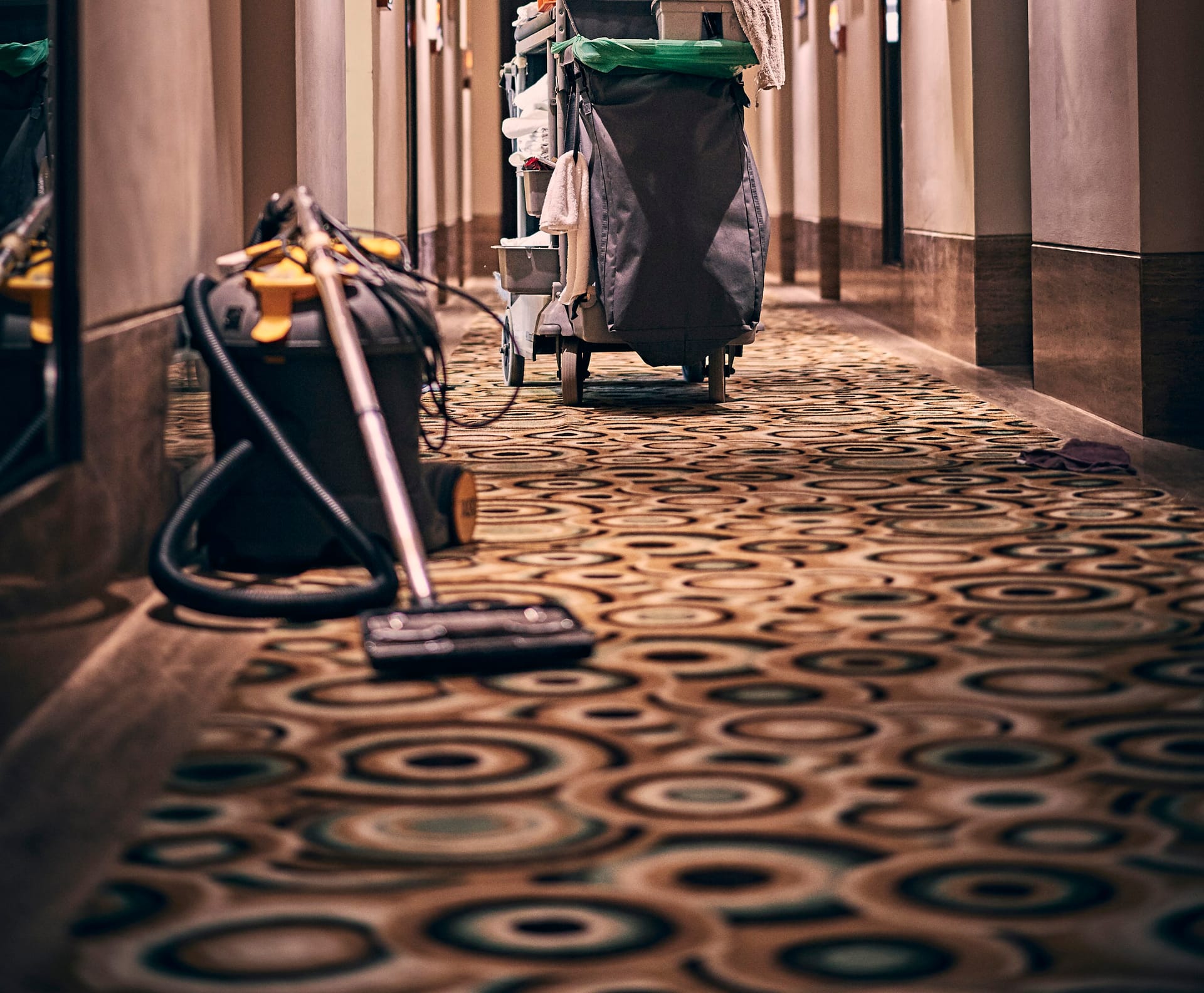 A commercial vacuum and housekeeping cart on the patterned carpet of a hotel corridor