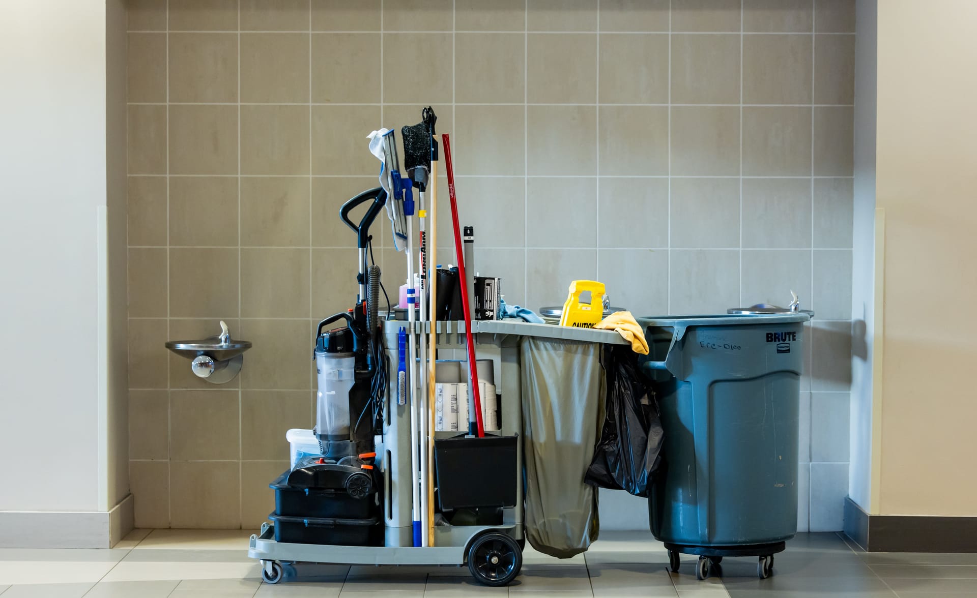 A stocked janitorial cart and waste bin parked in a commercial building corridor